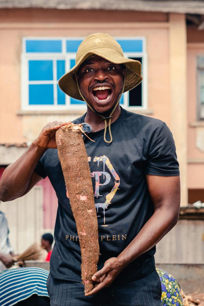 Smiling man holds a large cassava root outside a building, wearing a hat and T-shirt.