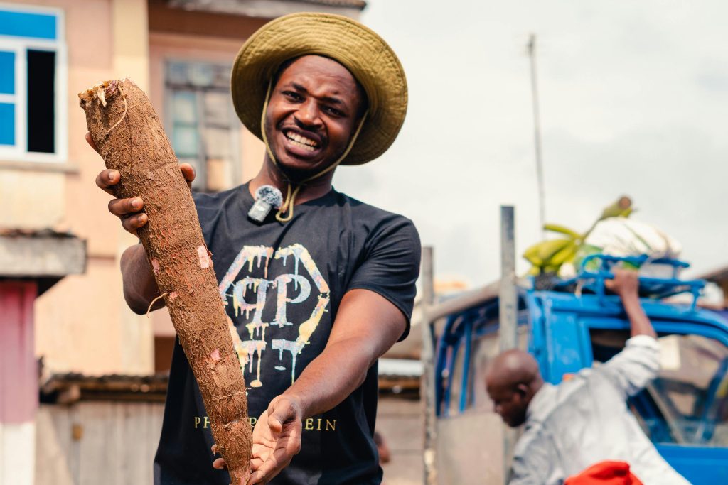 Smiling farmer proudly displays a large cassava root in an outdoor market setting.